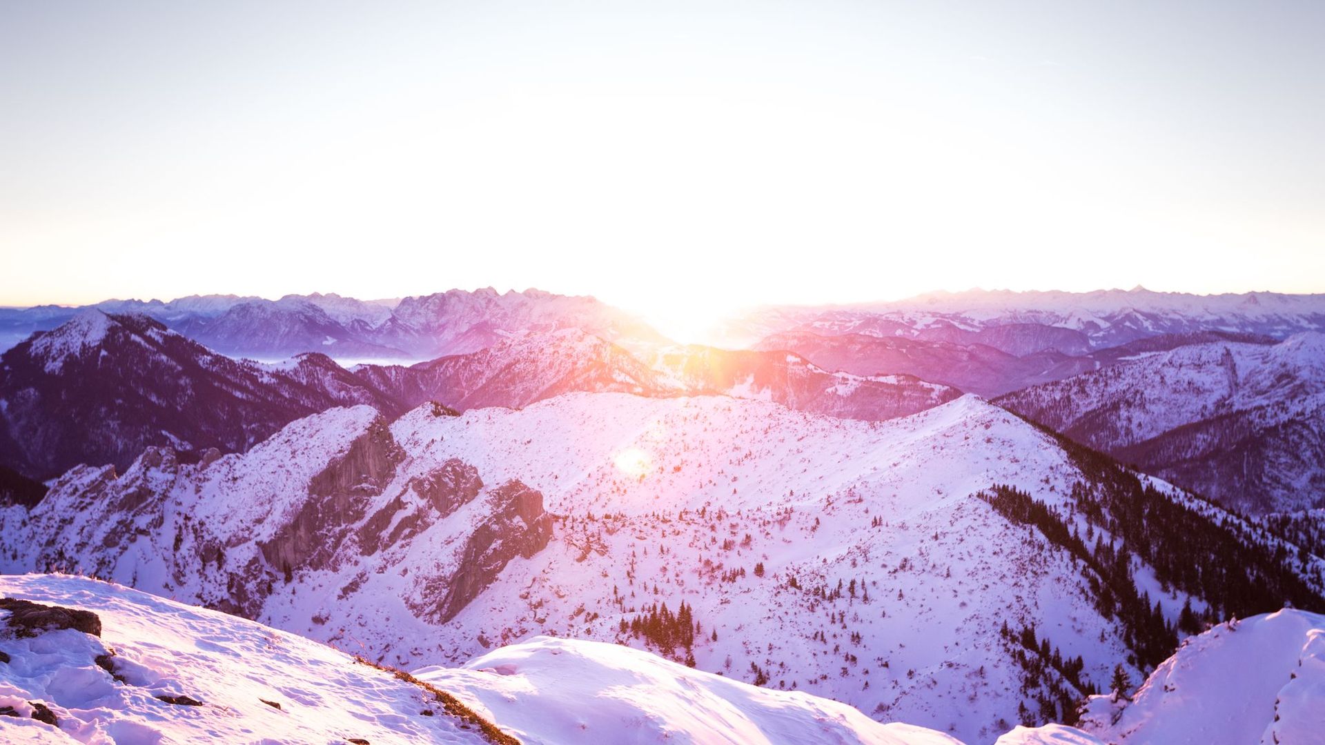 Sonnenaufgang über schneebedeckten Bergen mit sichtbaren Berggipfeln und vereinzelten Bäumen.