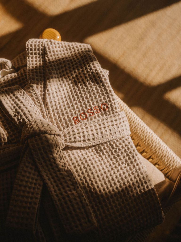 Ein gefalteter, beige waffle-texturierter Morgenmantel mit dem bestickten Schriftzug ‚ROSSO‘ liegt auf einem Stuhl aus hellen Holz- und Bastmaterialien, beleuchtet von warmem Sonnenlicht mit Schatten.