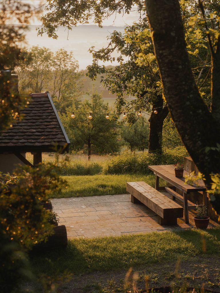 Gemütliche Sitzgelegenheit aus Holz mit Tisch und Bank auf einer gepflasterten Fläche im Garten, umgeben von Bäumen und mit warmem, spätnachmittäglichem Licht.