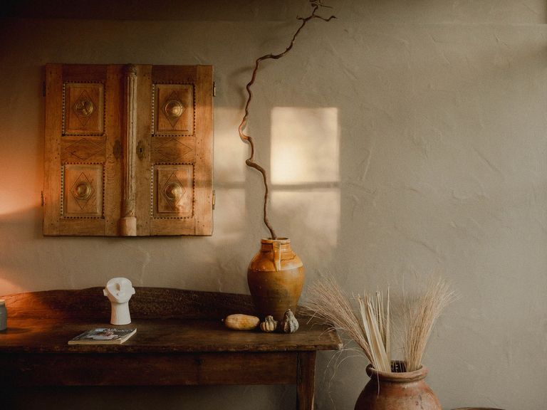 Ein rustikaler Holztisch vor einer beigen Wand mit einem alten, dekorativen hölzernen Fensterladen, einer Keramikvase mit einem langen, gebogenen Zweig, einigen kleinen Dekorationsgegenständen und einem großen Tonkrug mit getrockneten Gräsern.