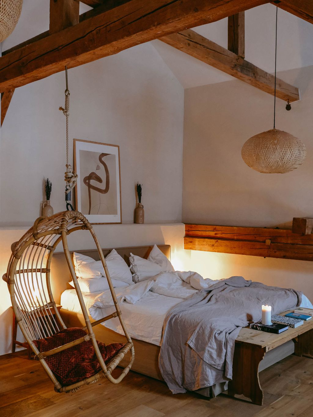 Gemütliches Schlafzimmer mit einem großen Bett mit weißen Kissen und Decken, einem an der Decke hängenden Korbsessel, rustikalen Holzbalken und warmem Licht.