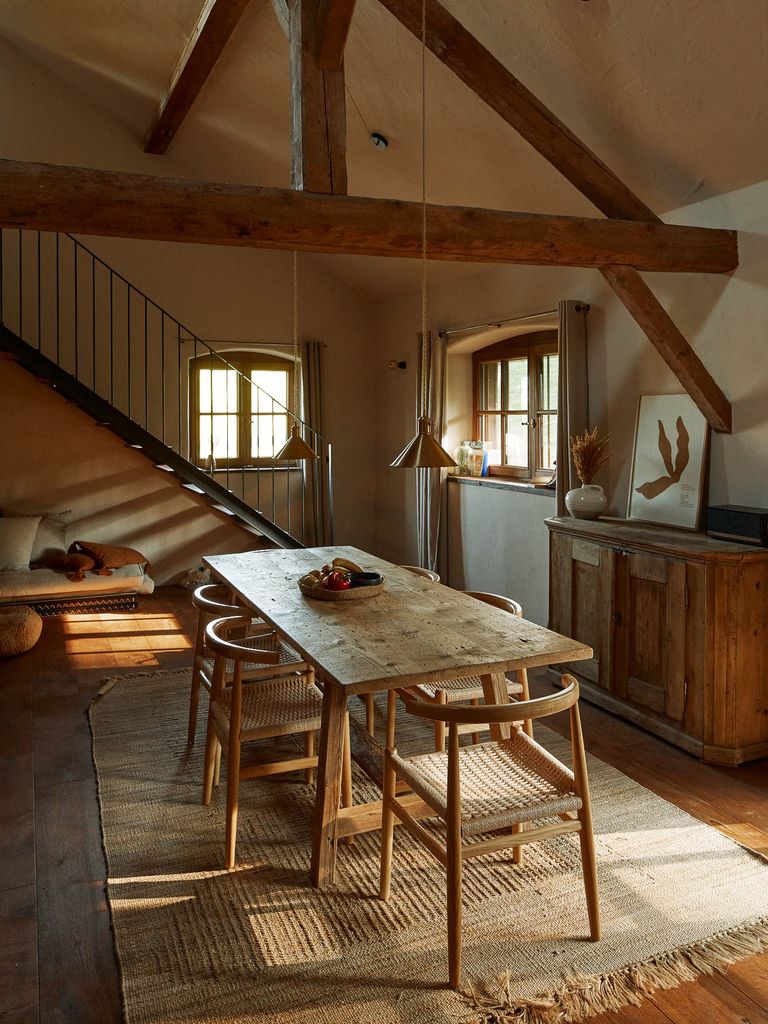 Rustikaler Essbereich mit hölzernem Tisch und sechs Stühlen auf einem Teppich, umgeben von sichtbaren Deckenbalken, einem Fenster mit Vorhängen und Kommode mit Dekoration.