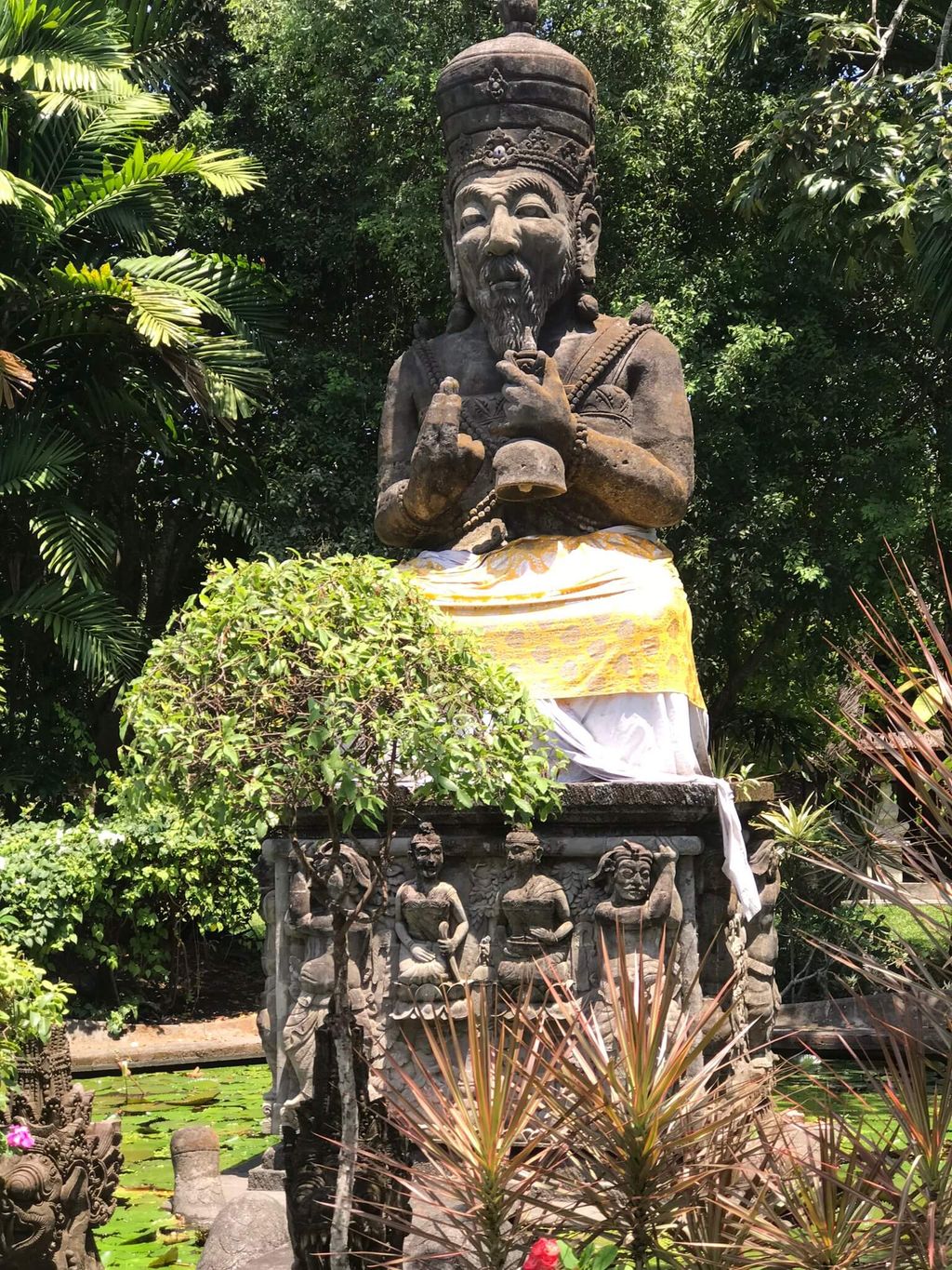 Stone statue of a seated man with traditional headwear surrounded by tropical plants in a garden in Bali.