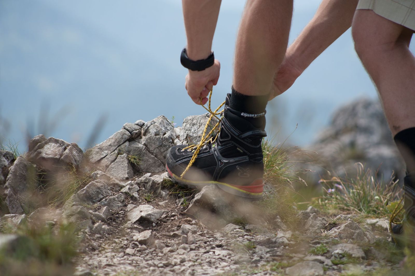 Ein Wanderer bindet seine Wanderschuhe auf felsigem Bergpfad mit Gras im Vordergrund.