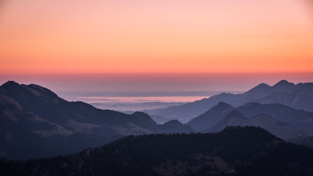 Blick auf eine Berglandschaft mit dunklen Konturen im Vordergrund und einem leuchtend orangefarbenen Himmel bei Sonnenaufgang oder Sonnenuntergang, im Hintergrund ist der Chiemsee zu sehen.