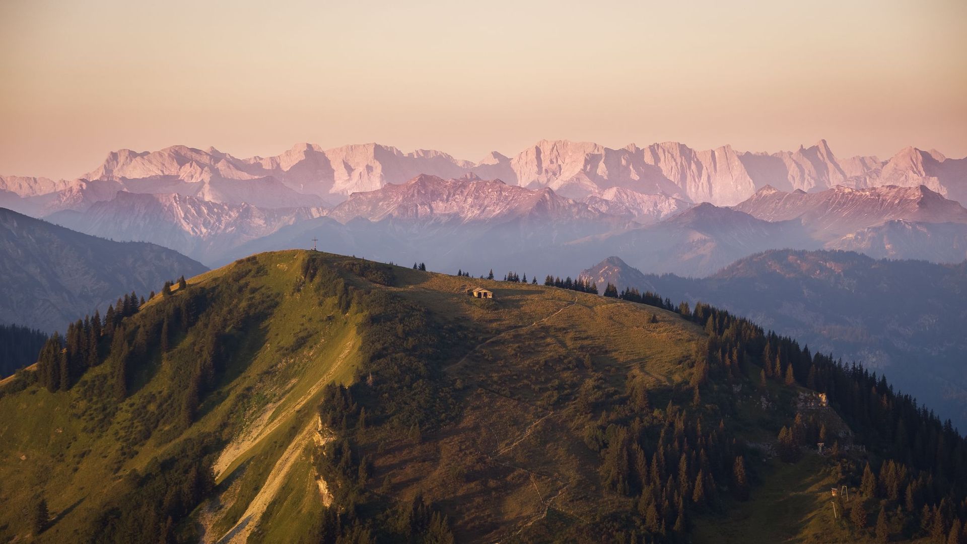 Blick auf einen bewaldeten Berggipfel im Vordergrund bei Sonnenuntergang mit den schneebedeckten Gipfeln des Karwendelgebirges im Hintergrund.