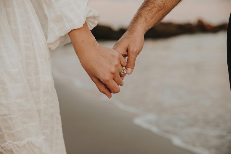 A couple holding hands on a beach with sand and water visible in the background.