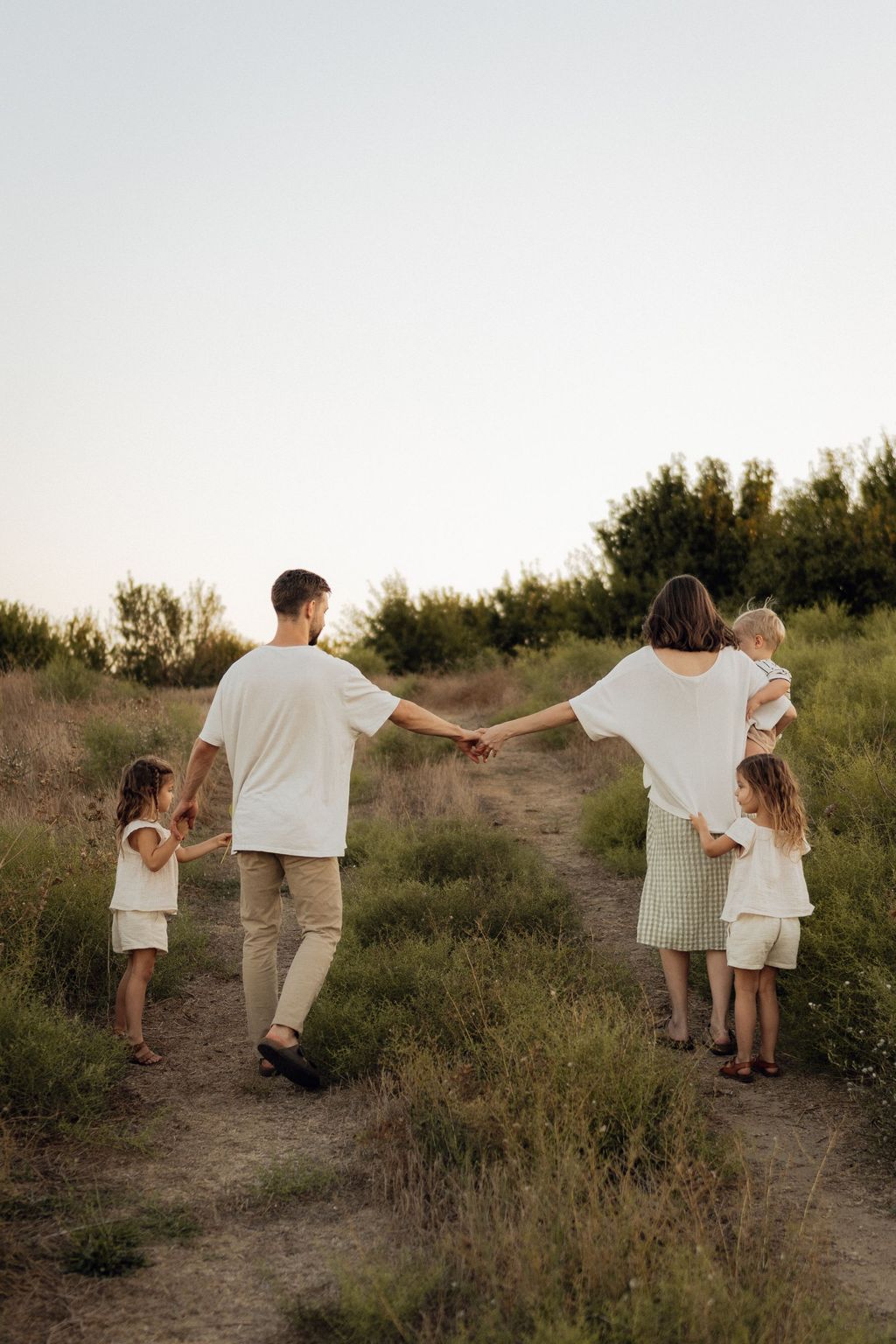 Eine Familie mit zwei Eltern und drei Kindern, die auf einem Weg durch eine natürliche, grasbewachsene Landschaft bei mildem Licht spazieren gehen, wobei die Eltern sich an den Händen halten.