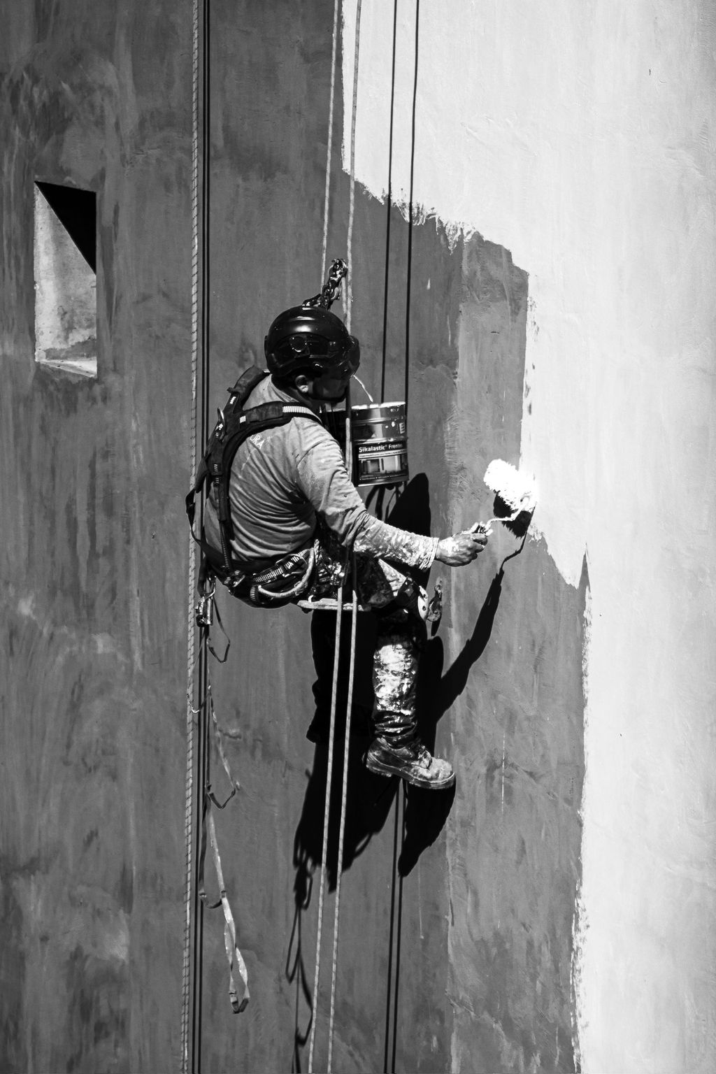 Ein Fassadenmaler mit Schutzhelm und Sicherheitsgeschirr streicht eine Wand in Weiß, während er an Seilen hängt.
