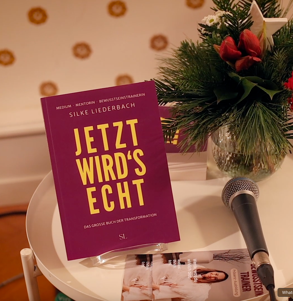 The book "Jetzt wird's echt" by Silke Liederbach is placed on a round white table next to a microphone and floral decoration.