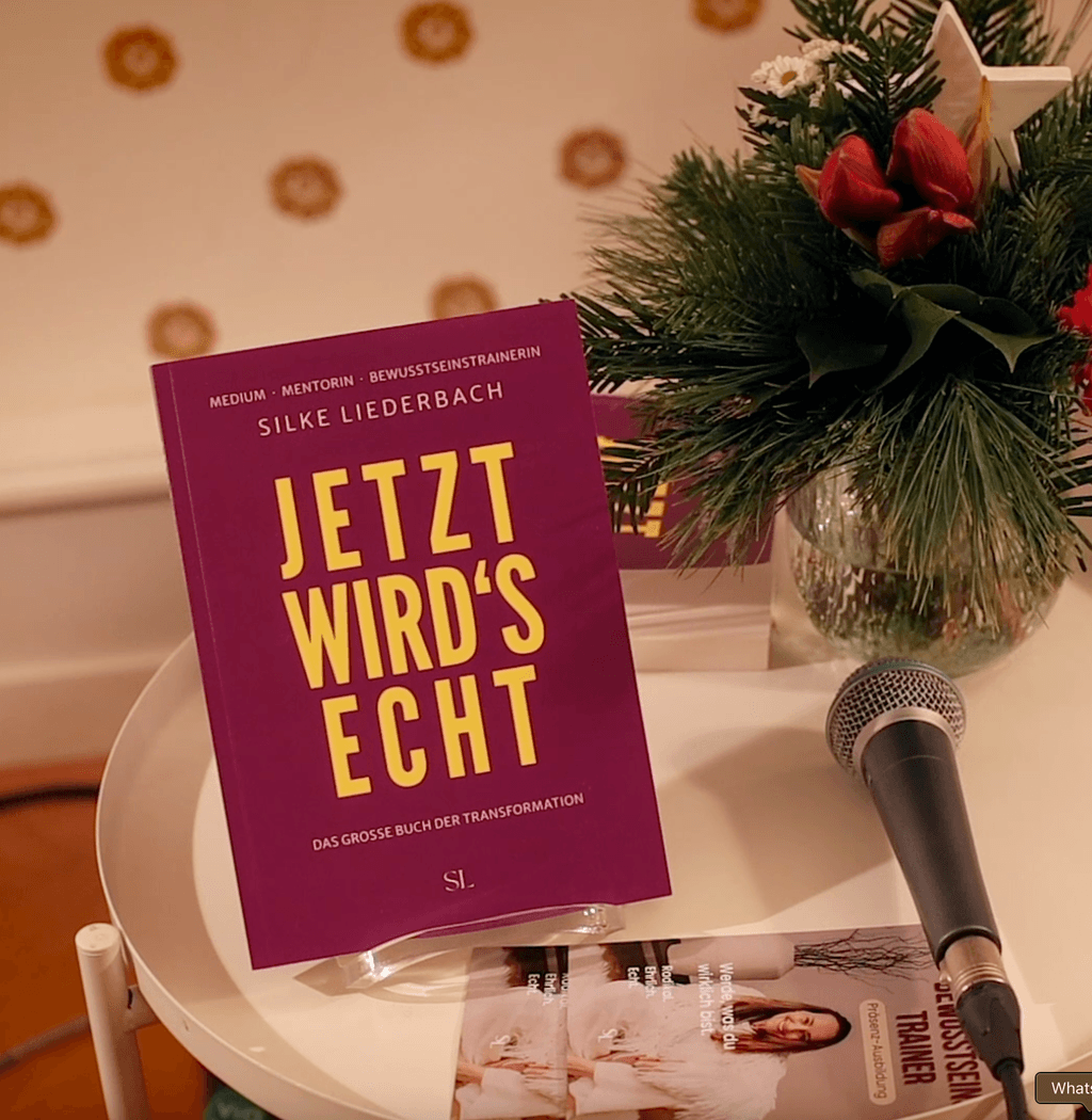Das Buch "Jetzt wird's echt" von Silke Liederbach steht auf einem weißen runden Tisch neben einem Mikrofon und einem kleinen Blumenarrangement mit roten Blüten und grünen Nadeln.