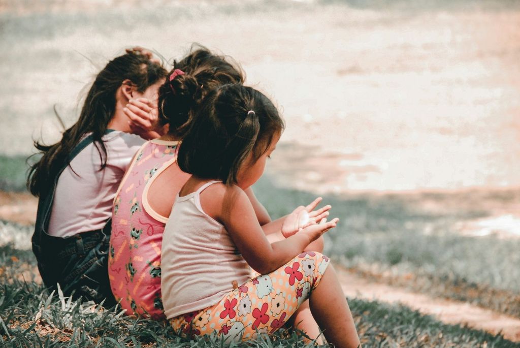 Three young children sitting on grass facing away, with one child in a white tank top and floral shorts, another in a pink dress with cartoon prints, and the third in a light blue shirt with suspenders.