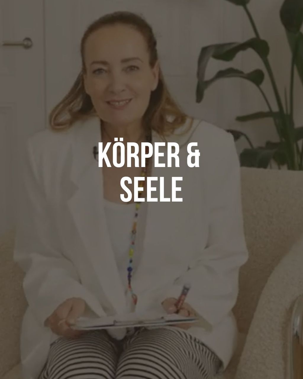 Woman sitting on a sofa holding a clipboard and writing, with a houseplant in the background. The text 'Körper & Seele' is overlaid.
