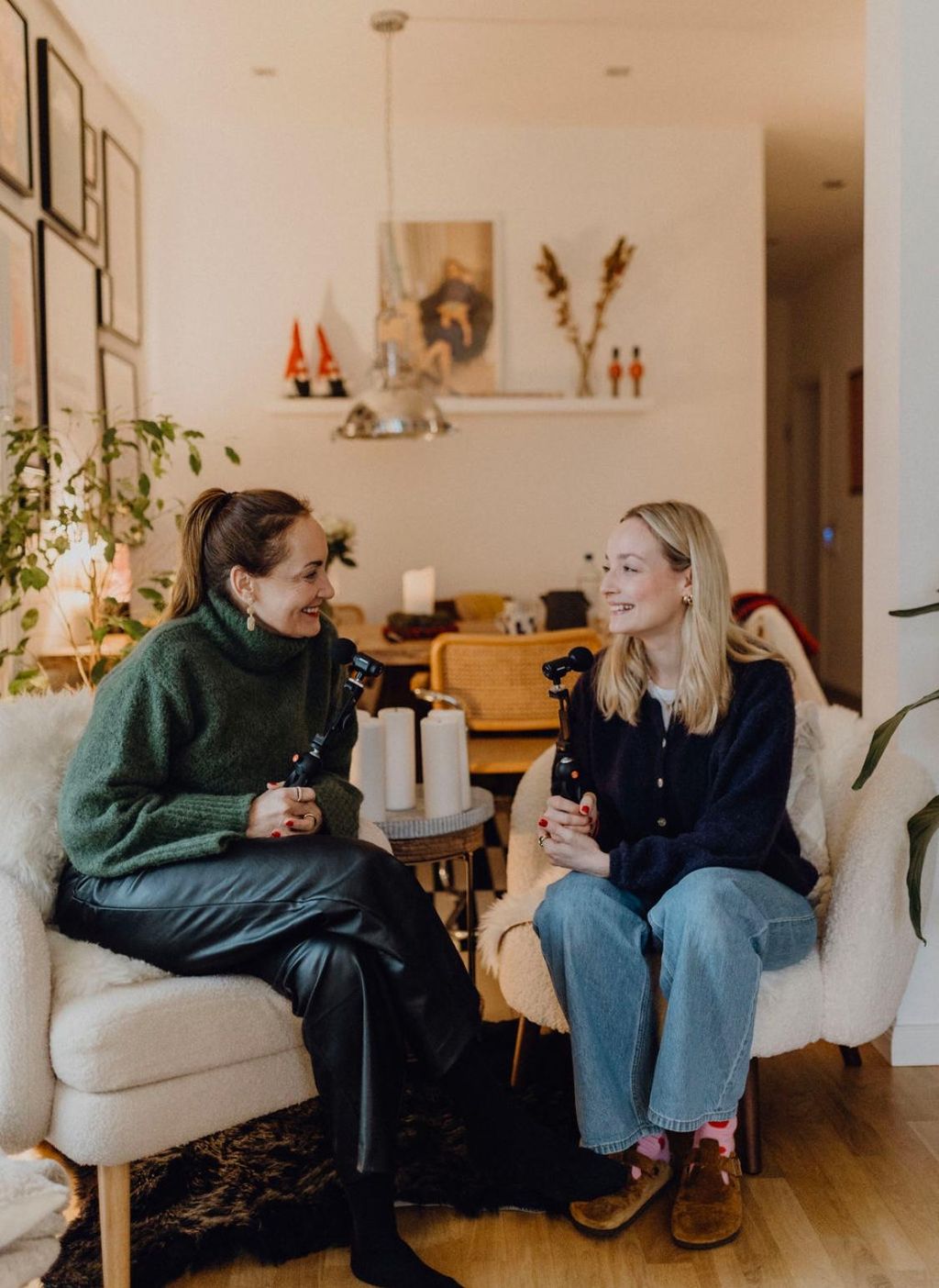 Two women sit in a modern, cozy living room on armchairs holding microphones while talking and smiling at each other.