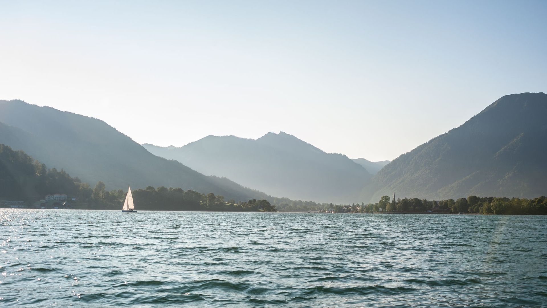 Segelboot mit weißem Segel auf einem ruhigen See vor einer Kulisse von bewaldeten Bergen unter klarem Himmel.
