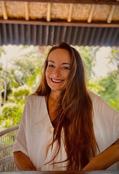 Eine lächelnde Frau mit langen braunen Haaren, die in einer weißen Bluse auf einer Veranda mit natürlicher und grüner Umgebung sitzt.