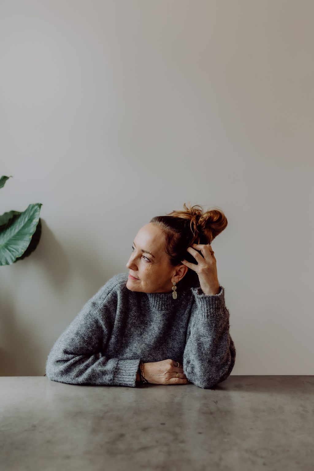 Frau mit hochgestecktem Haar und grauem Pullover sitzt an einem Tisch und schaut nach links an eine helle Wand.