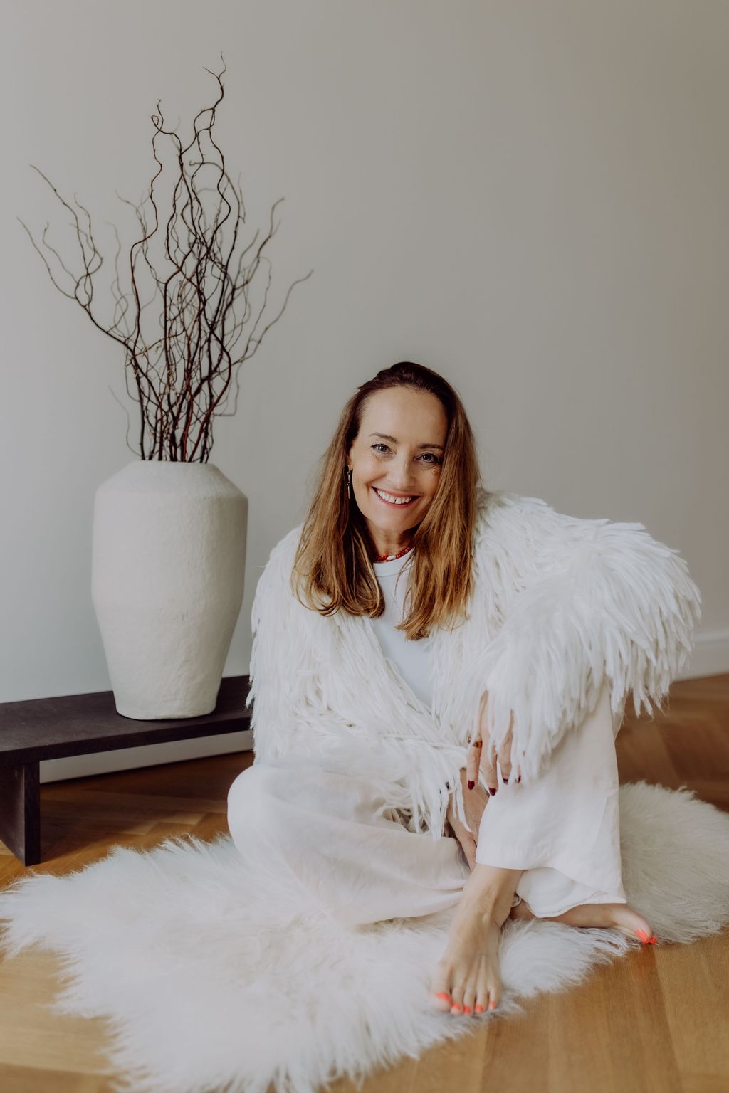Eine lächelnde Frau mit langem braunem Haar sitzt auf einem weißen Fellteppich auf einem Holzboden neben einer dunkelbraunen Bank mit einer großen weißen Vase voller dünner Äste.