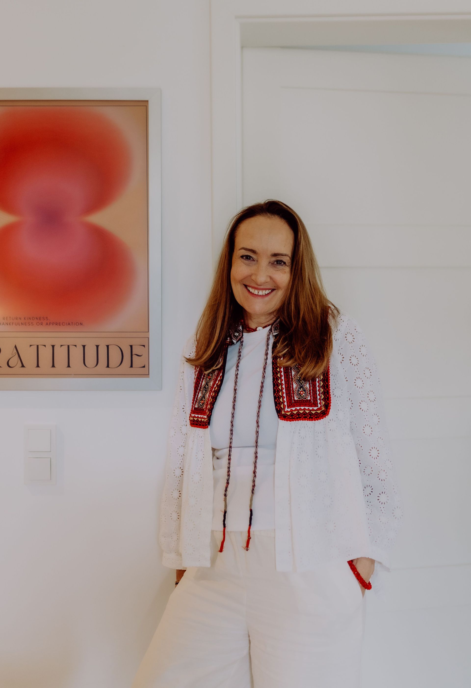 Eine lächelnde Frau mit langen braunen Haaren steht vor einer weißen Tür neben einem Wandbild mit dem Wort 'GRATITUDE'. Sie trägt eine weiße Bluse mit roten und schwarzen Stickereien.
