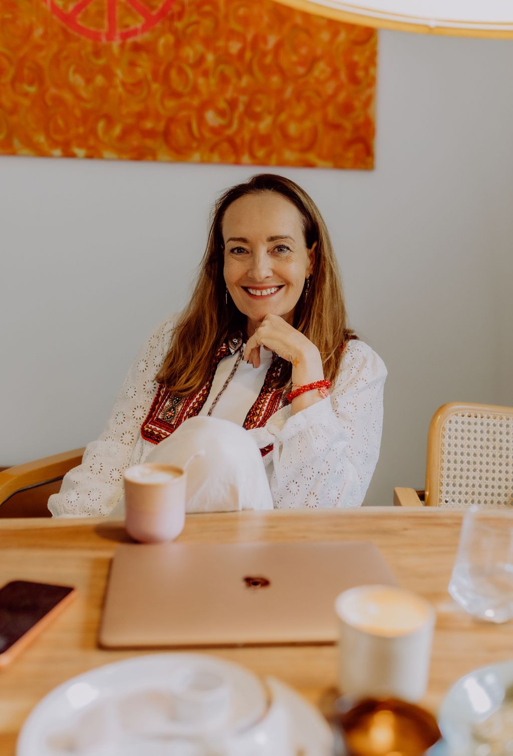A woman in white clothing with a red embroidered detail sits relaxed at a wooden table with a laptop, cup, and glasses in a modern room with an orange wall painting.