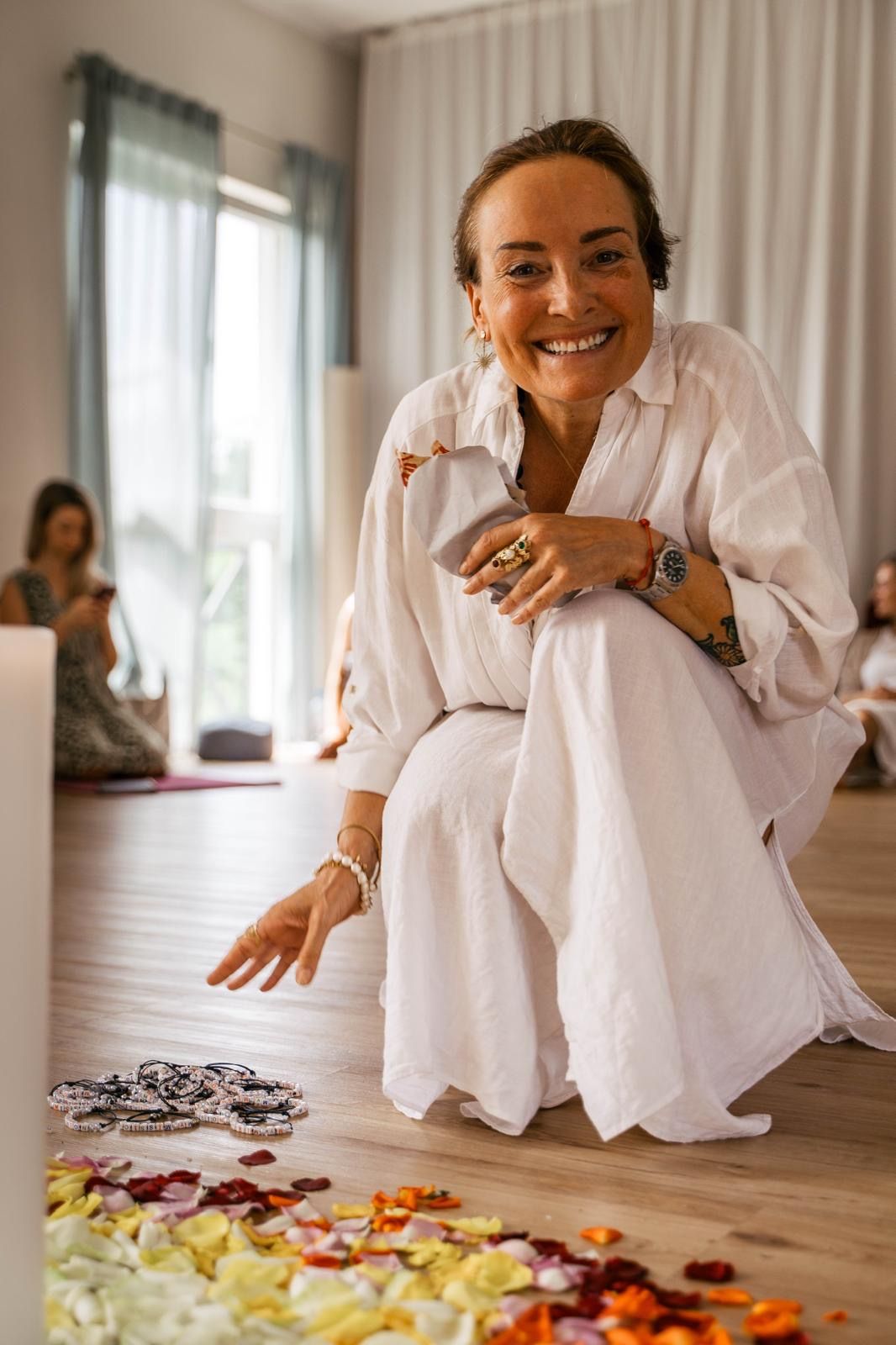 Eine lächelnde Frau in weißem Kleid hockt in einem hellen Raum auf dem Boden und hält eine Stoffserviette, vor ihr liegen bunte Blütenblätter und Ketten auf dem Holzboden.