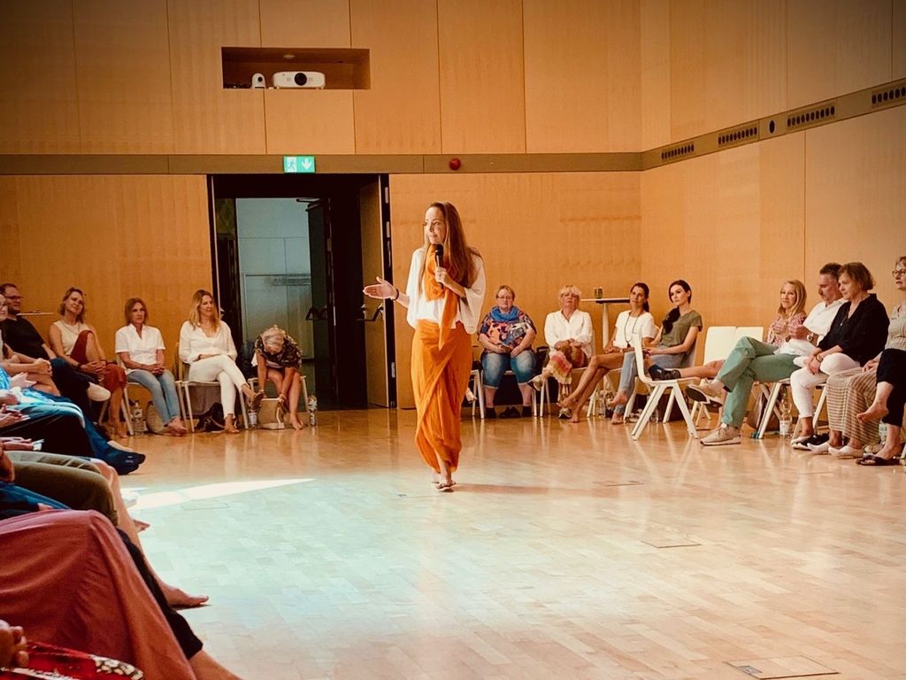 Eine Frau hält ein Live-Seminar in einem großen Raum mit Holzverkleidung, während ein Publikum auf Stühlen rundherum sitzt.