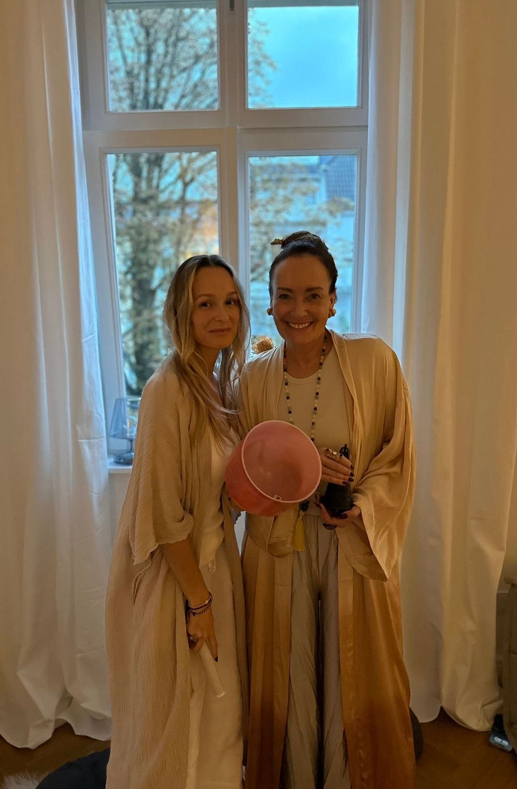 Two women in light robes stand in front of a window with white curtains, one holding a pink singing bowl and the other a white stick.