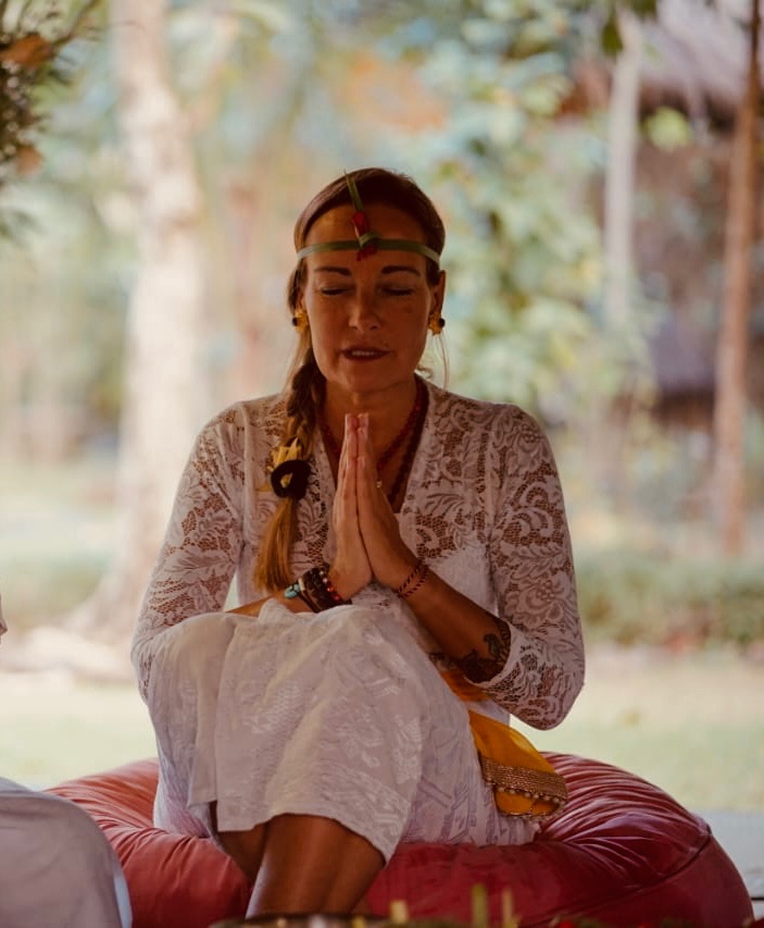 Woman in white lace clothing sitting outdoors on a red cushion with closed eyes and hands pressed together in prayer.