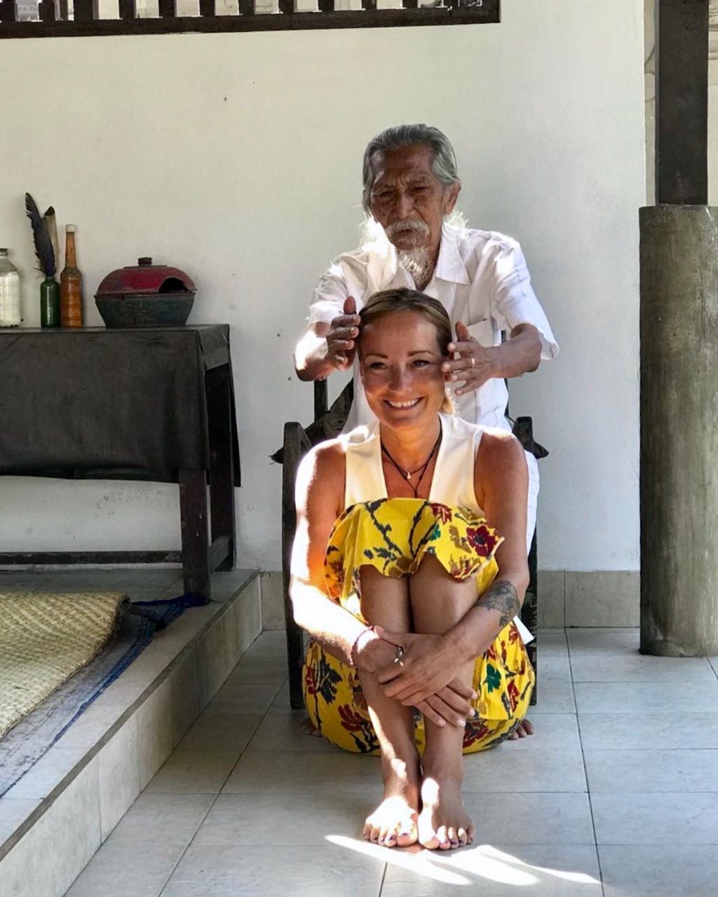 An older man holding his hands over the head of a smiling woman sitting on the floor during a healing ceremony in a bright room.