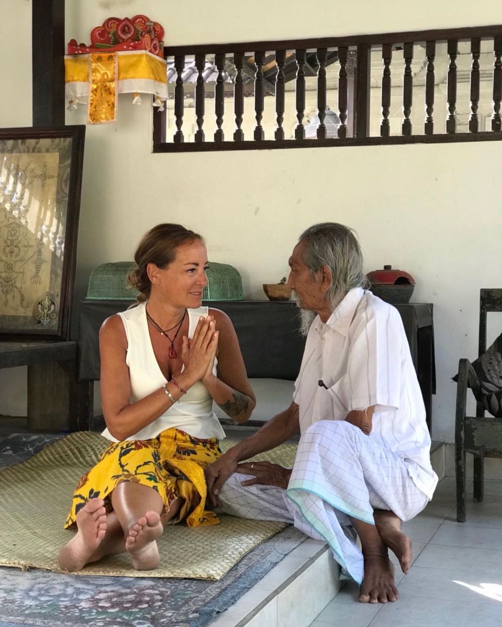 A woman and an elderly man sit opposite each other on a mat on the floor in a room with traditional decorations, engaged in a friendly and respectful conversation.