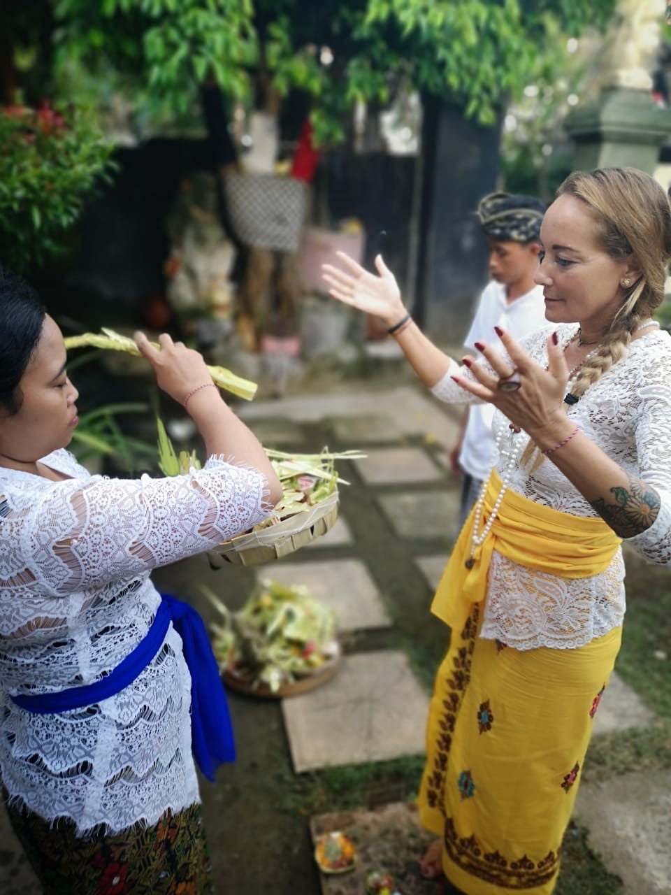 Two women and a boy in traditional Balinese attire participating in an outdoor ceremony, one woman holding a basket of offerings.