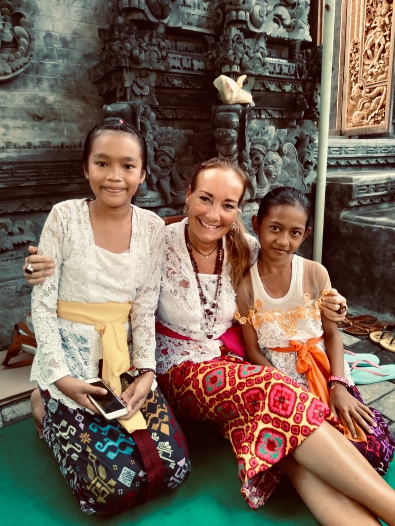 Three women wearing traditional Balinese clothing sit on a green mat in front of an intricately carved stone relief.
