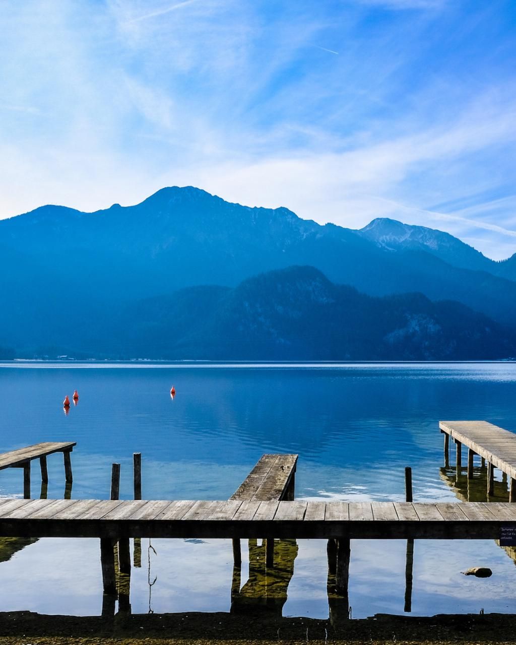 Wooden pier at the calm Tegernsee lake with reflective water and mountain backdrop under a blue sky.