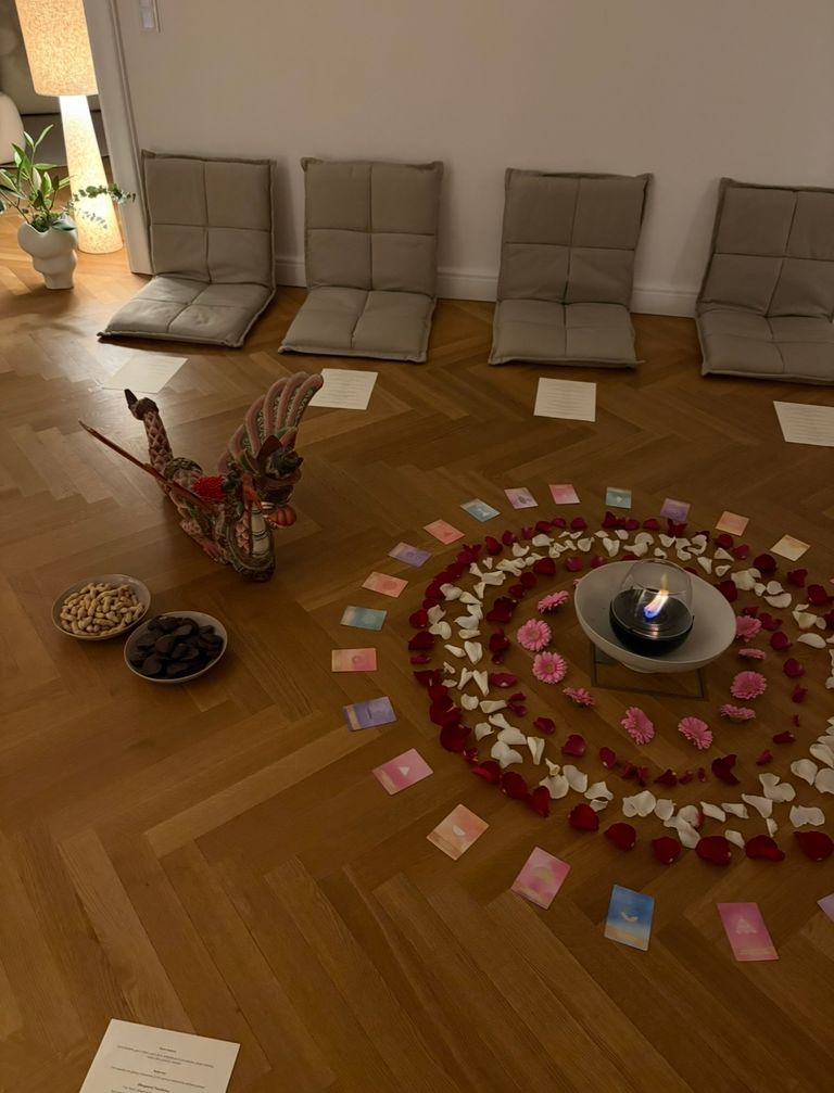 A circle of colorful cards, rose petals, and pink flowers surrounds a burning candle on a wooden floor, with four comfortable floor cushions and a decorative dragon figure nearby.