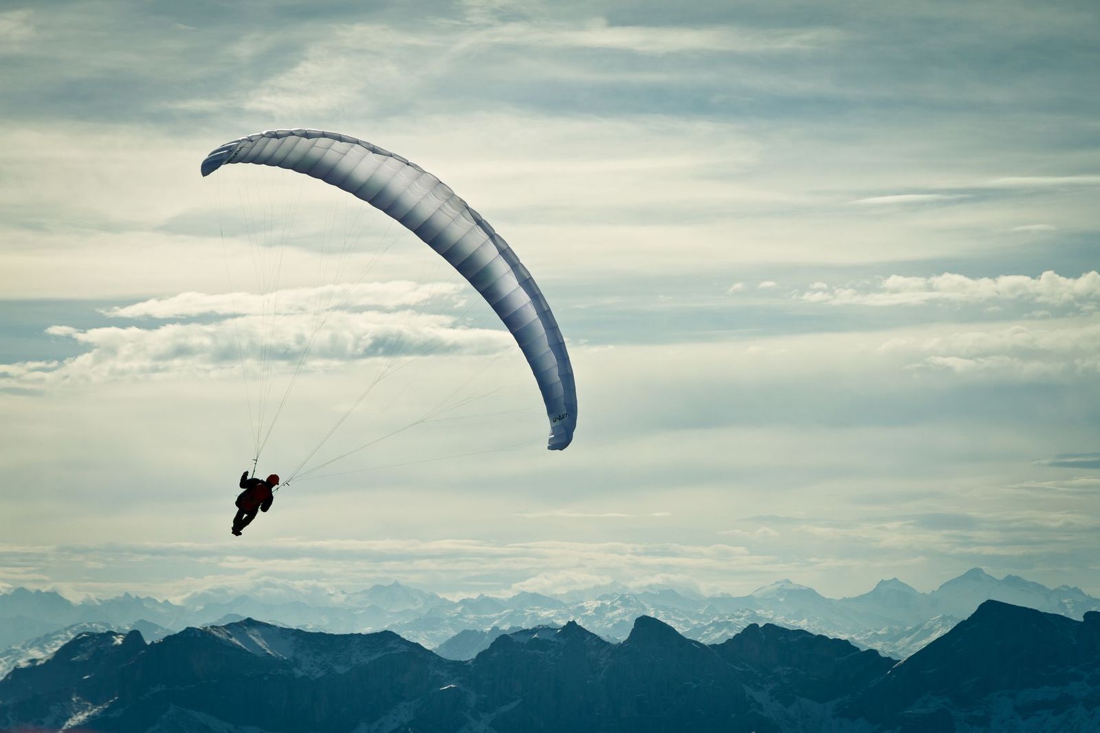 Person mit Gleitschirm, die über Berglandschaft mit bewölktem Himmel gleitet.